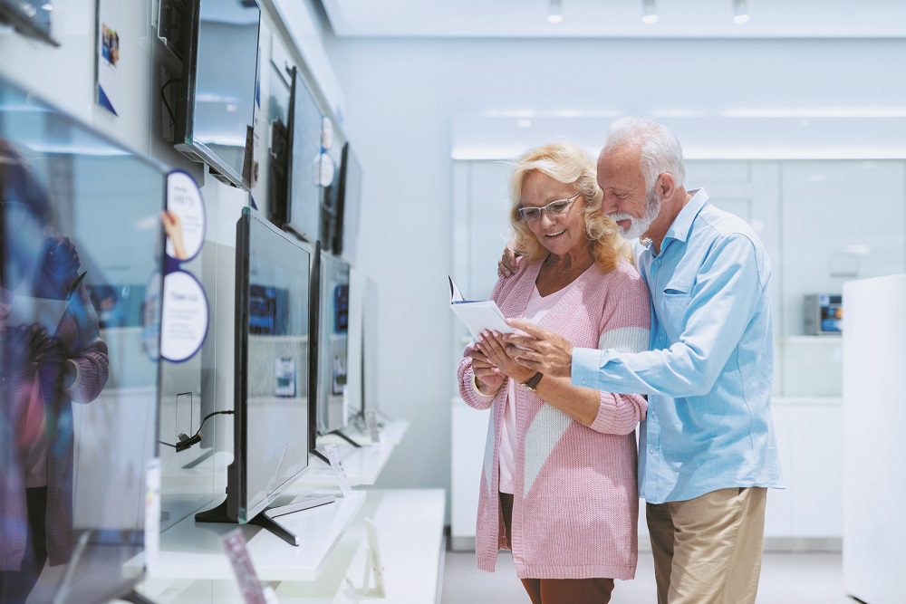 Cute smiling Caucasian senior couple standing in tech store and looking at prices for tv they want to buy.