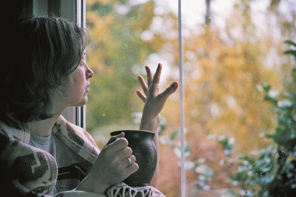Fall melancholy mood concept. Wistful woman sitting near window and looking at autumn landscape outside in rainy day. Copy space.