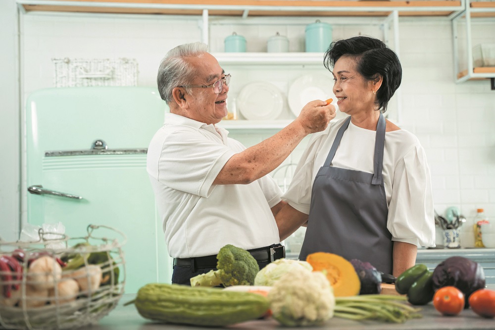Senior Asian couple grandparents cooking together while woman is feeding food to man at the kitchen. Long lasting relationship concept